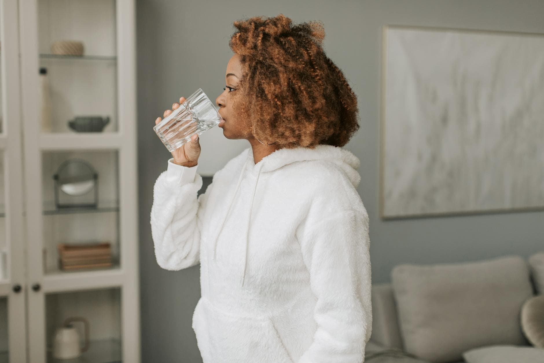 A Black woman with beautiful natural hair holds a glass of water, symbolizing the importance of hydration for healthy hair growth, set in a bright and inviting environment.